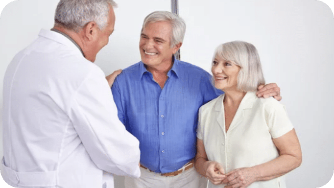 Eine fachkundige Person im weißen Kittel spricht freundlich mit einem älteren Paar. Alle drei lächeln, das Paar steht nah beieinander und wirkt entspannt. Die Szene vermittelt ein wertschätzendes, unterstützendes Gespräch.