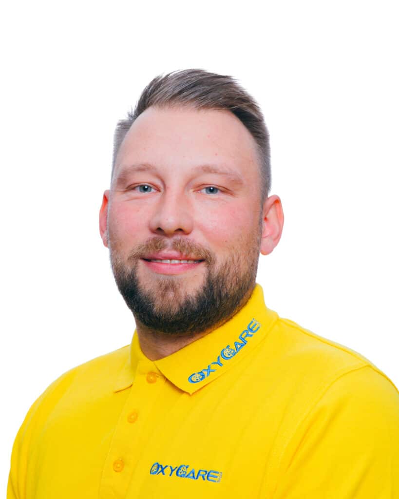 Foto von Dennis Bühring, Mitarbeiter von OxyCare. Er trägt ein gelbes Hemd mit dem OxyCare Logo und schaut freundlich in die Kamera.
