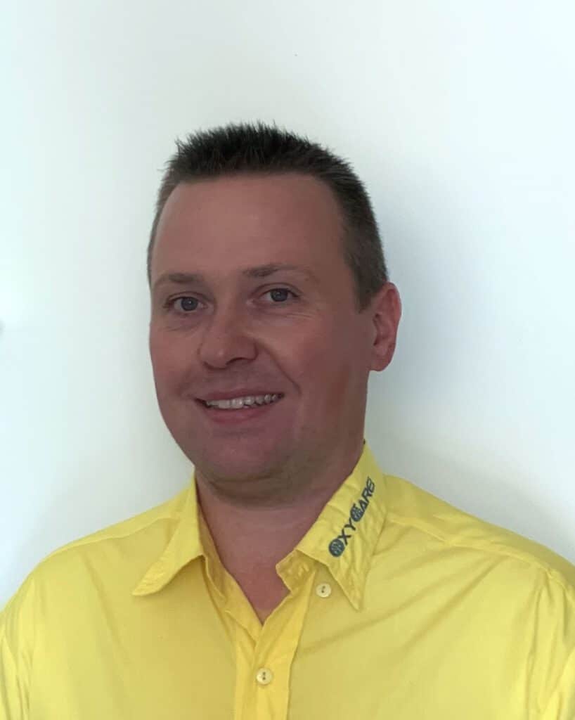 Foto von Benjamin Alberding, Mitarbeiter von OxyCare. Er trägt ein gelbes Hemd mit dem OxyCare Logo und schaut freundlich in die Kamera.
