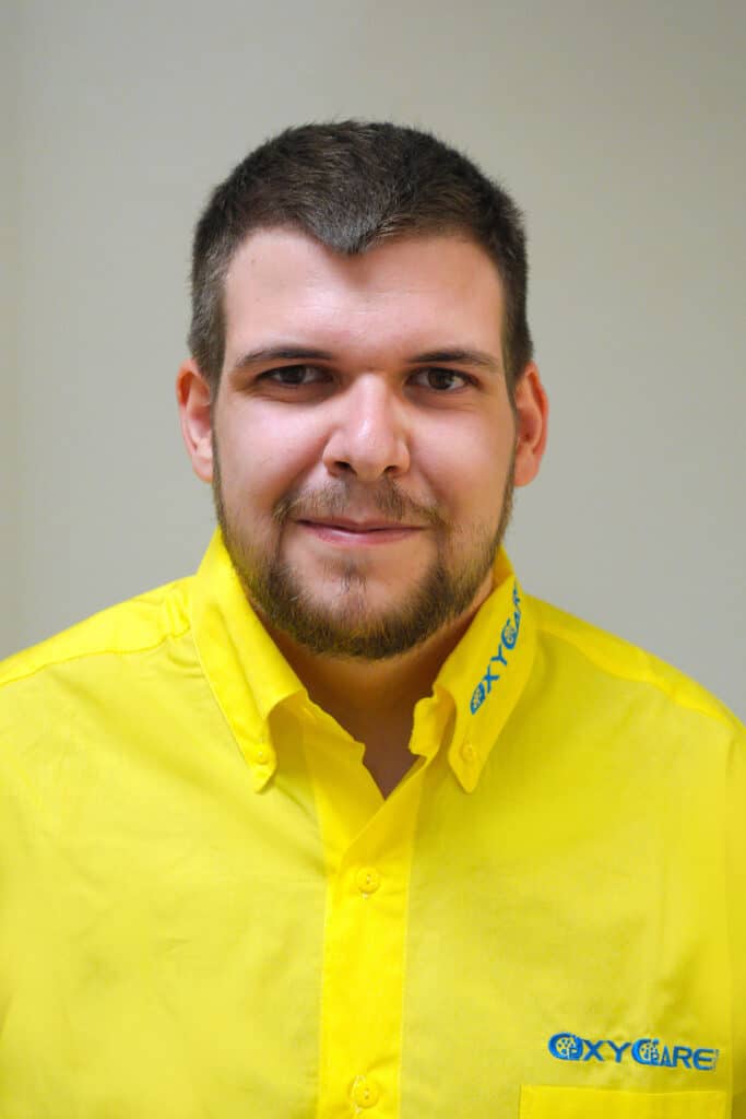 Foto von Alexander Burger, Mitarbeiter von OxyCare. Er trägt ein gelbes Hemd mit dem OxyCare Logo und schaut freundlich in die Kamera.