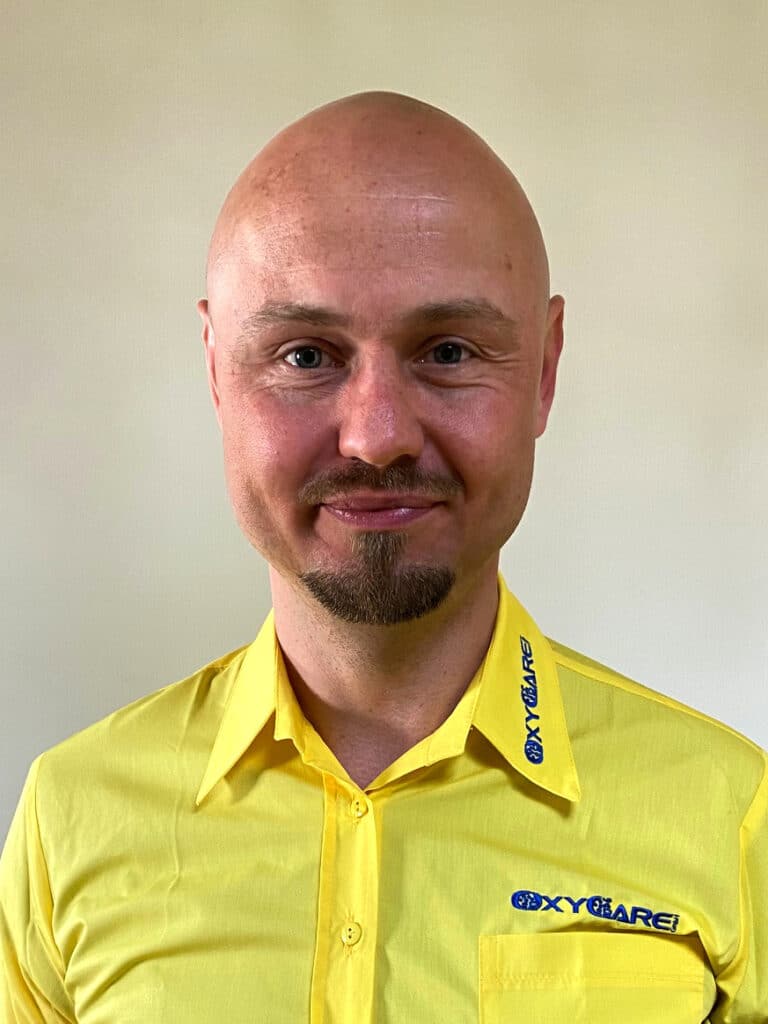 Foto von Denis Bastijan, Mitarbeiter von OxyCare. Er trägt ein gelbes Hemd mit dem OxyCare Logo und schaut freundlich in die Kamera.