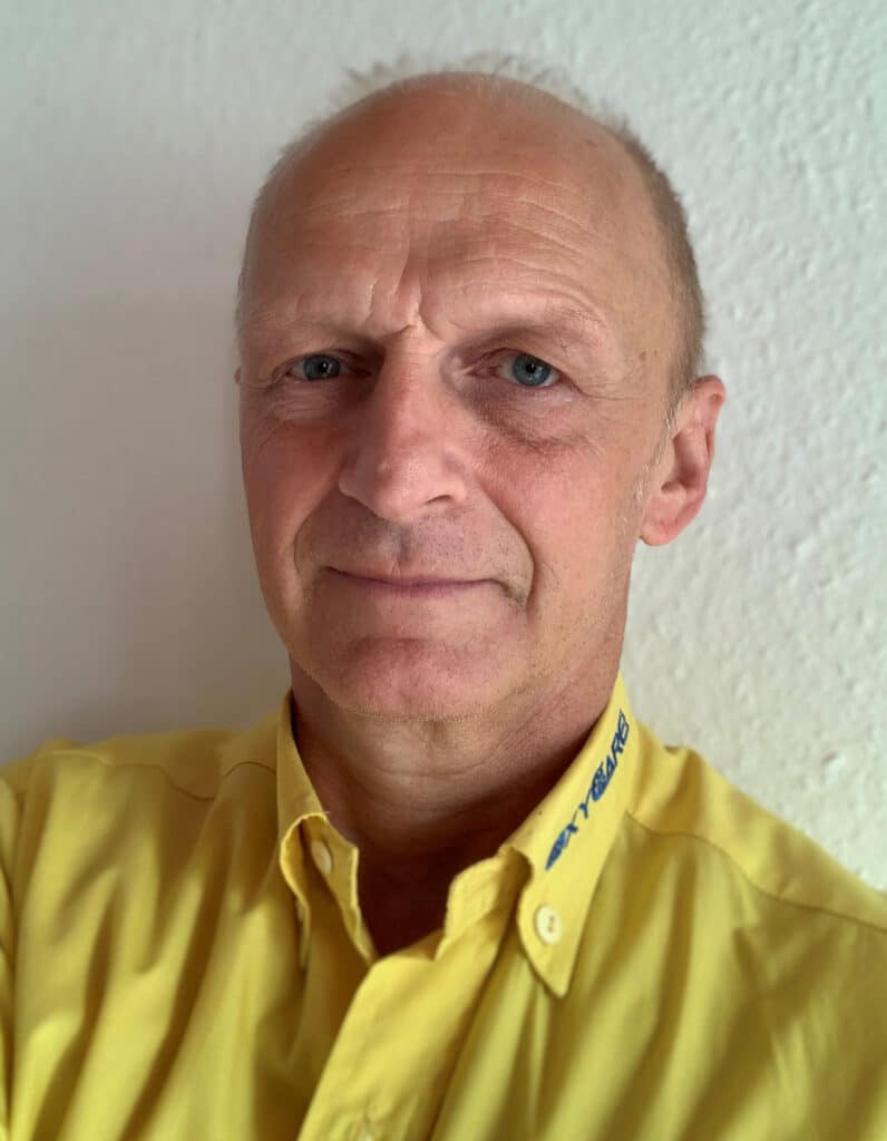 Foto von Gerhard Bender, Mitarbeiter von OxyCare. Er trägt ein gelbes Hemd mit dem OxyCare Logo und schaut freundlich in die Kamera.