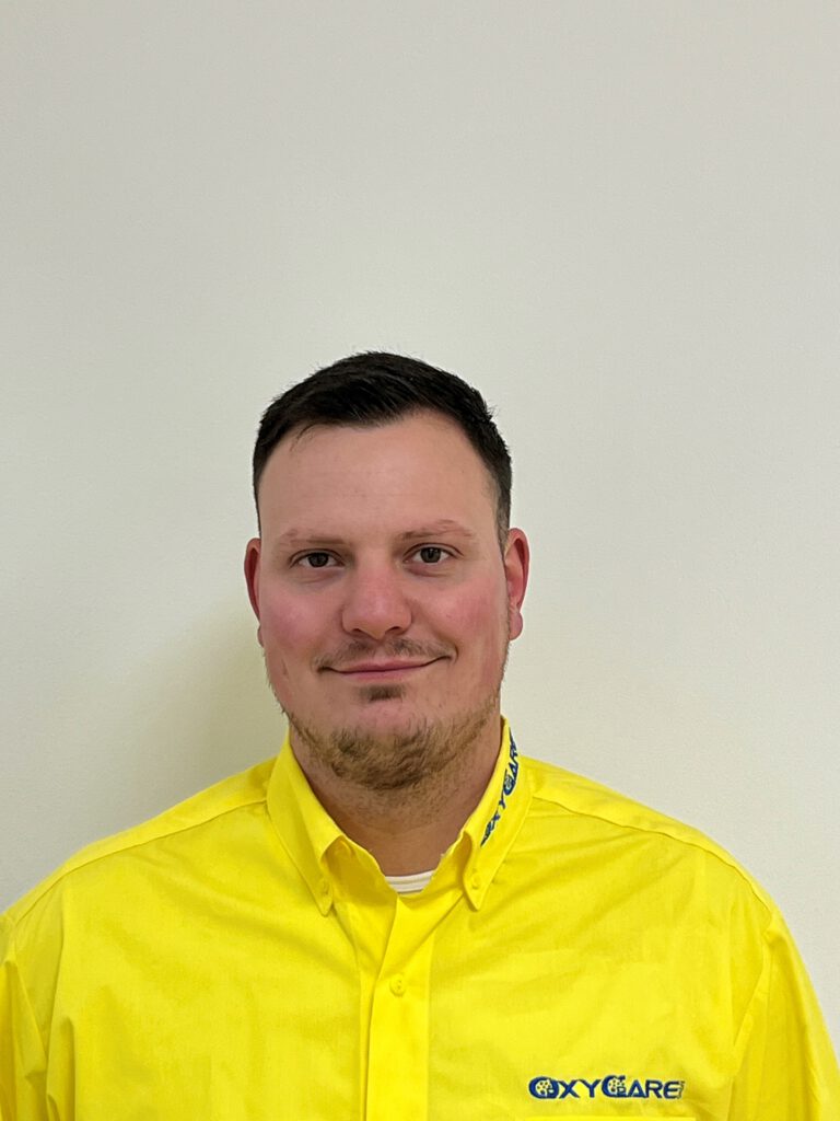 Foto von Tobias Ehlert, Mitarbeiter von OxyCare. Er trägt ein gelbes Hemd mit dem OxyCare Logo und schaut freundlich in die Kamera.