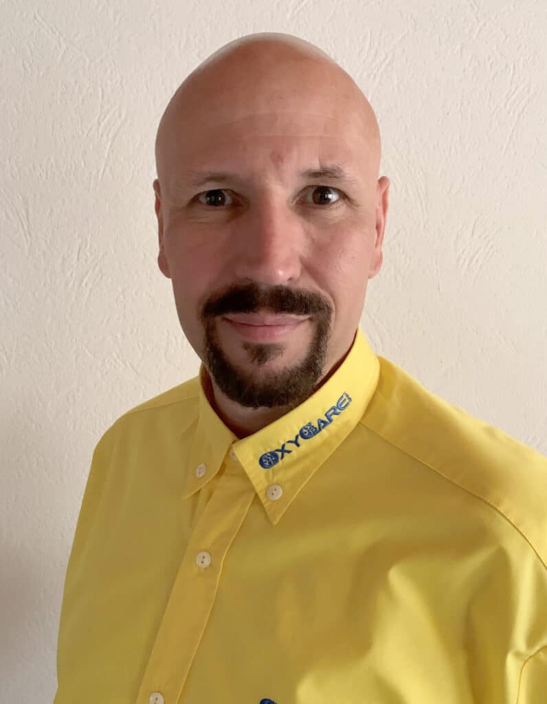 Foto von Gunther Grimm, Mitarbeiter von OxyCare. Er trägt ein gelbes Hemd mit dem OxyCare Logo und schaut freundlich in die Kamera.