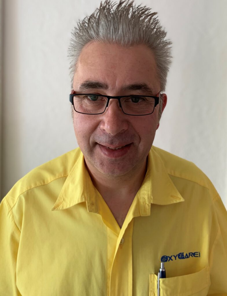 Foto von Oliver Grüter, Mitarbeiter von OxyCare. Er trägt ein gelbes Hemd mit dem OxyCare Logo und schaut freundlich in die Kamera.