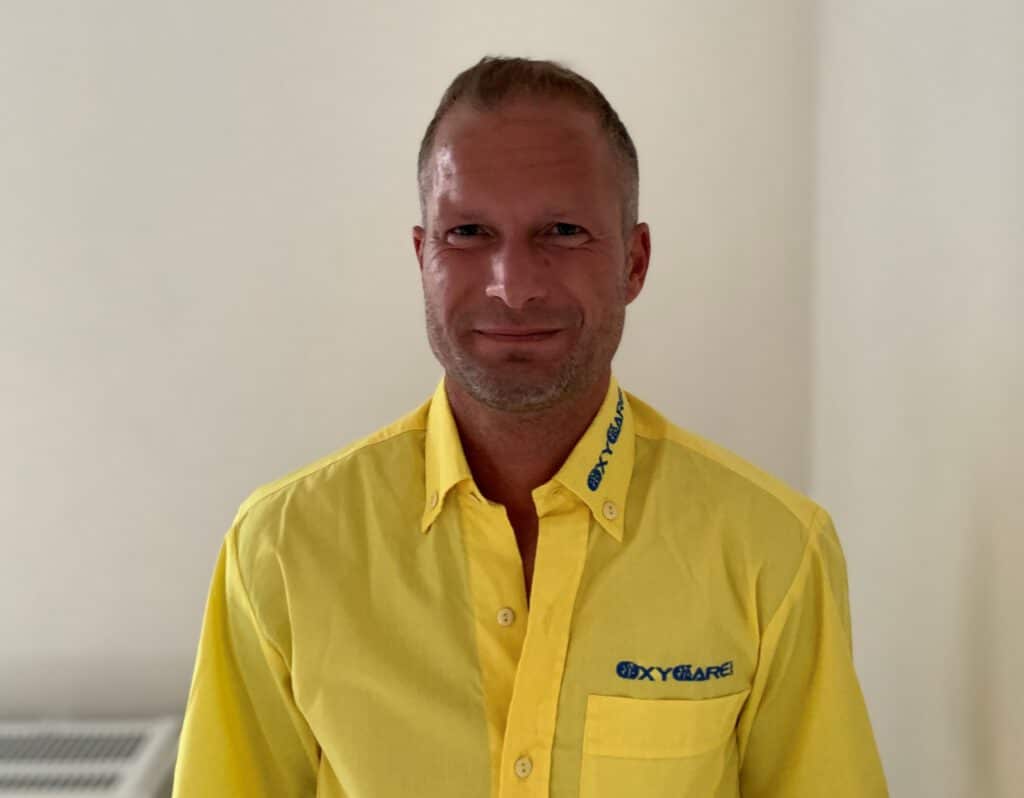 Foto von Benjamin Hartung, Mitarbeiter von OxyCare. Er trägt ein gelbes Hemd mit dem OxyCare Logo und schaut freundlich in die Kamera.