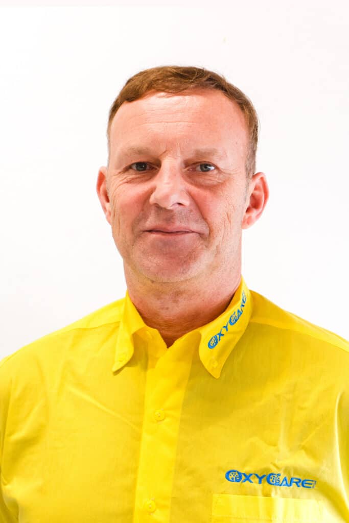 Foto von Heiko Leistner, Mitarbeiter von OxyCare. Er trägt ein gelbes Hemd mit dem OxyCare Logo und schaut freundlich in die Kamera.