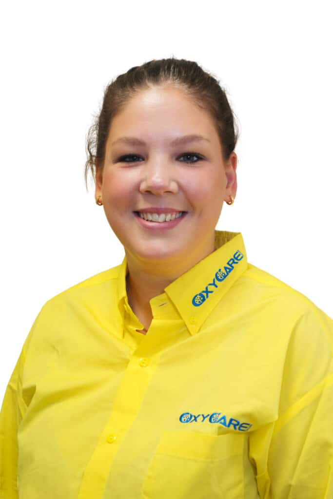Foto von Alina Janike Ramadani, Mitarbeiterin von OxyCare. Sie trägt ein gelbes Hemd mit dem OxyCare Logo und schaut freundlich in die Kamera.