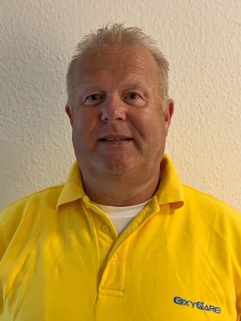 Foto von Gunnar John, Mitarbeiter von OxyCare. Er trägt ein gelbes Hemd mit dem OxyCare Logo und schaut freundlich in die Kamera.