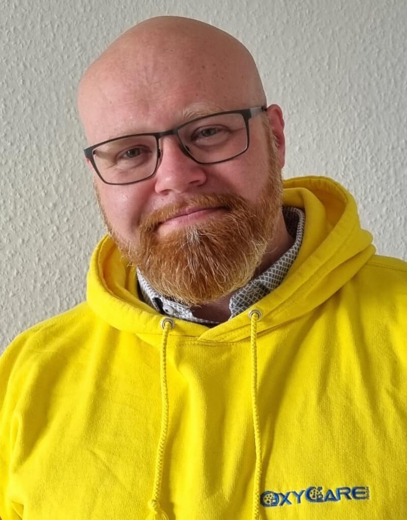 Foto von Keith Neitzel, Mitarbeiter von OxyCare. Er trägt einen gelben Pullover mit dem OxyCare Logo und schaut freundlich in die Kamera.