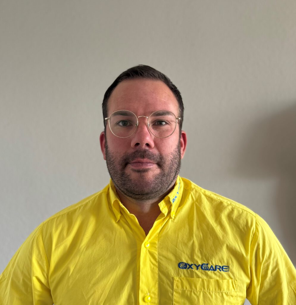 Foto von Christopher Strupat, Mitarbeiter von OxyCare. Er trägt ein gelbes Hemd mit dem OxyCare Logo und schaut freundlich in die Kamera.