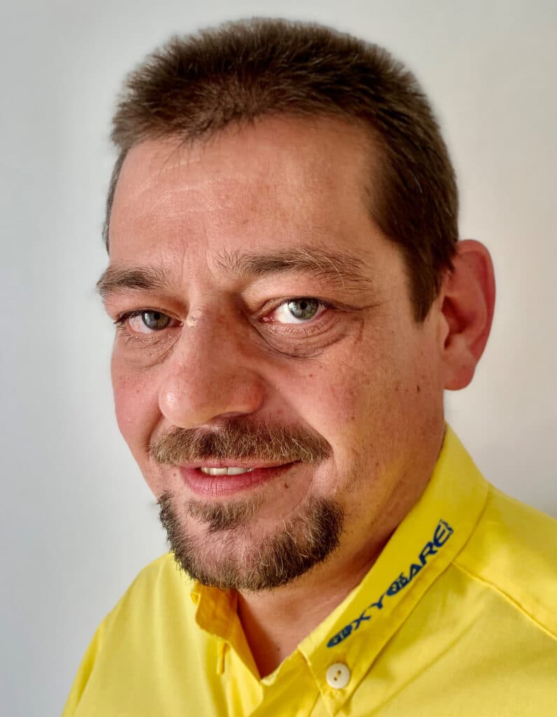 Foto von Carlheinz Veit, Mitarbeiter von OxyCare. Er trägt ein gelbes Hemd mit dem OxyCare Logo und schaut freundlich in die Kamera.