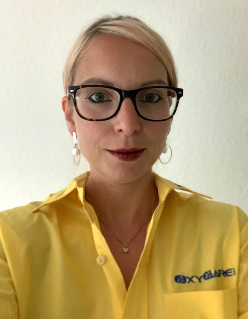 Foto von Dorothee Wolf, Mitarbeiterin von OxyCare. Sie trägt ein gelbes Hemd mit dem OxyCare Logo und schaut freundlich in die Kamera.