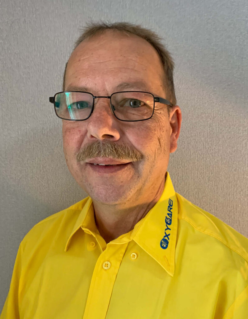 Foto von Carsten Axthelm, Mitarbeiter von OxyCare. Er trägt ein gelbes Hemd mit dem OxyCare Logo und schaut freundlich in die Kamera.
