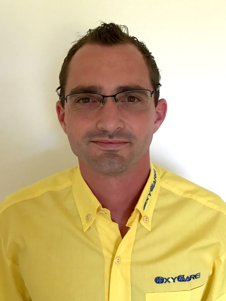 Foto von Christopher Bohrer, Mitarbeiter von OxyCare. Er trägt ein gelbes Hemd mit dem OxyCare Logo und schaut freundlich in die Kamera.