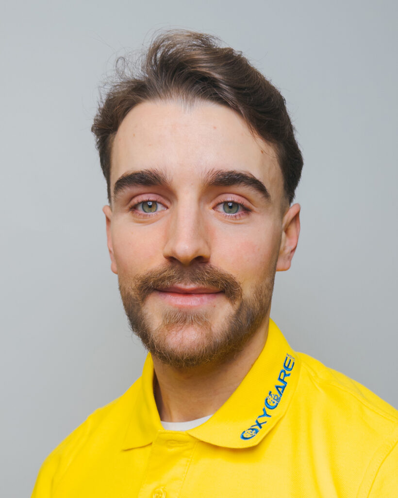 Foto von Eliot Muriqi, Mitarbeiter von OxyCare. Er trägt ein gelbes Hemd mit dem OxyCare Logo und schaut freundlich in die Kamera.