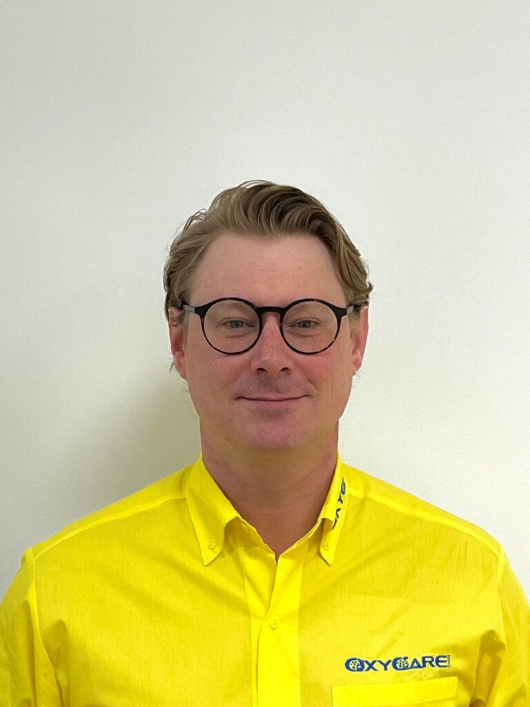 Foto von Gero Frese, Mitarbeiter von OxyCare. Er trägt ein gelbes Hemd mit dem OxyCare Logo und schaut freundlich in die Kamera.