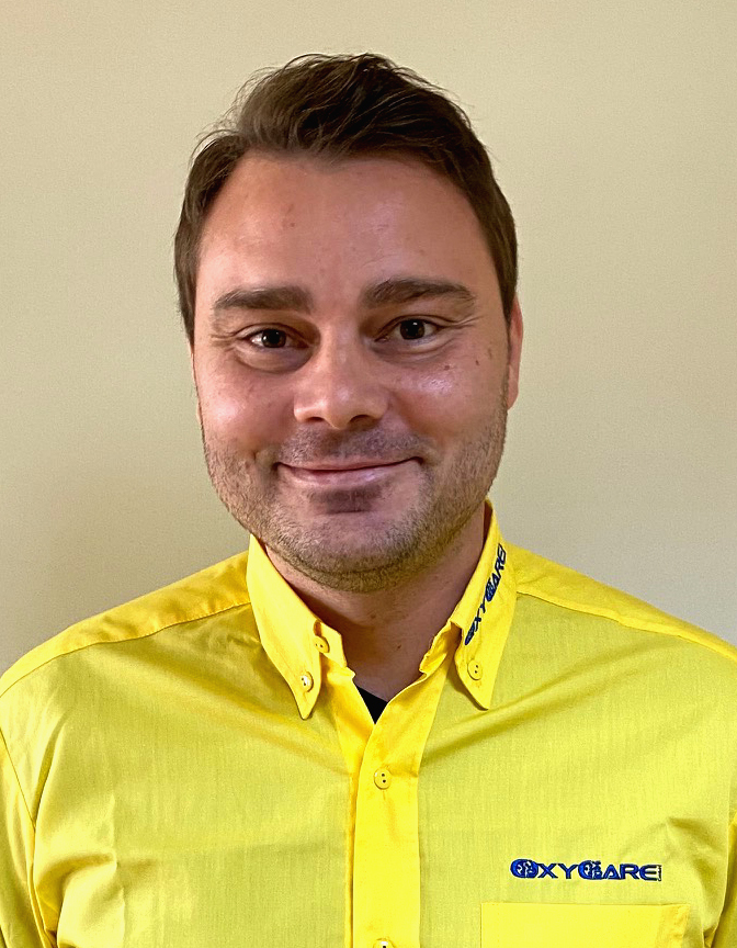 Foto von Sergei Jakob, Mitarbeiter von OxyCare. Er trägt ein gelbes Hemd mit dem OxyCare Logo und schaut freundlich in die Kamera.