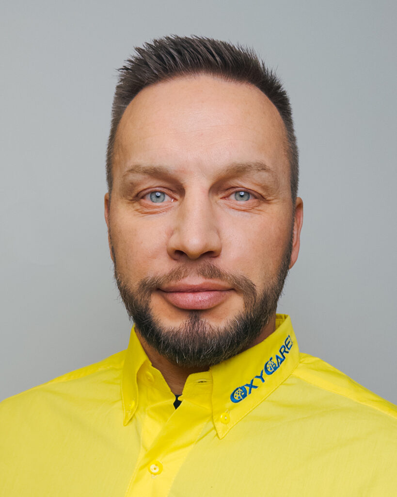 Foto von Nico Kasbaum, Mitarbeiter von OxyCare. Er trägt ein gelbes Hemd mit dem OxyCare Logo und schaut freundlich in die Kamera.