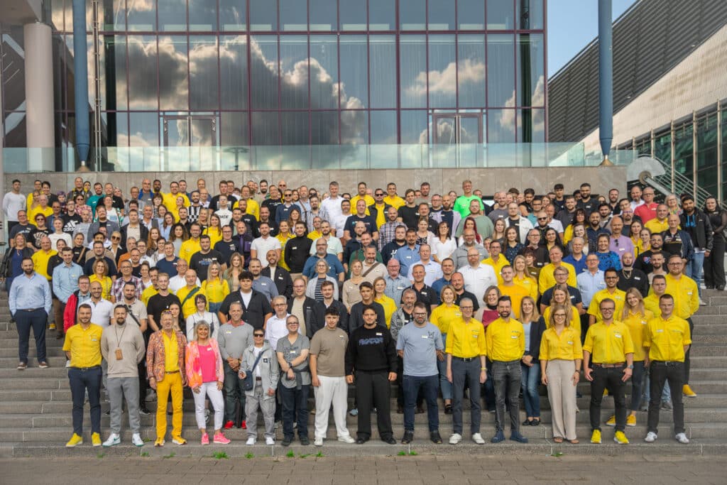 Großes Gruppenfoto mit mehreren hundert OxyCare Mitarbeitenden auf einer Treppe vor einem modernen Gebäude. Viele tragen gelbe OxyCare-Oberteile. Die Menschen stehen dicht beieinander, lächeln und schauen in die Kamera. Das Bild zeigt die Teilnehmenden der großen OxyCare Schulung 2024 in Bremen.