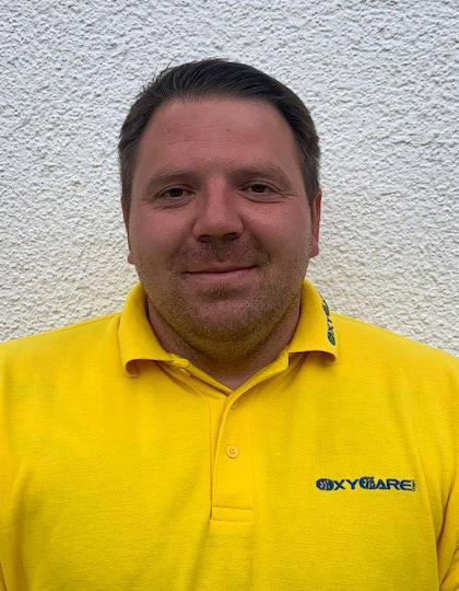 Foto von Marco Schreiner, Mitarbeiter von OxyCare. Er trägt ein gelbes Hemd mit dem OxyCare Logo und schaut freundlich in die Kamera.