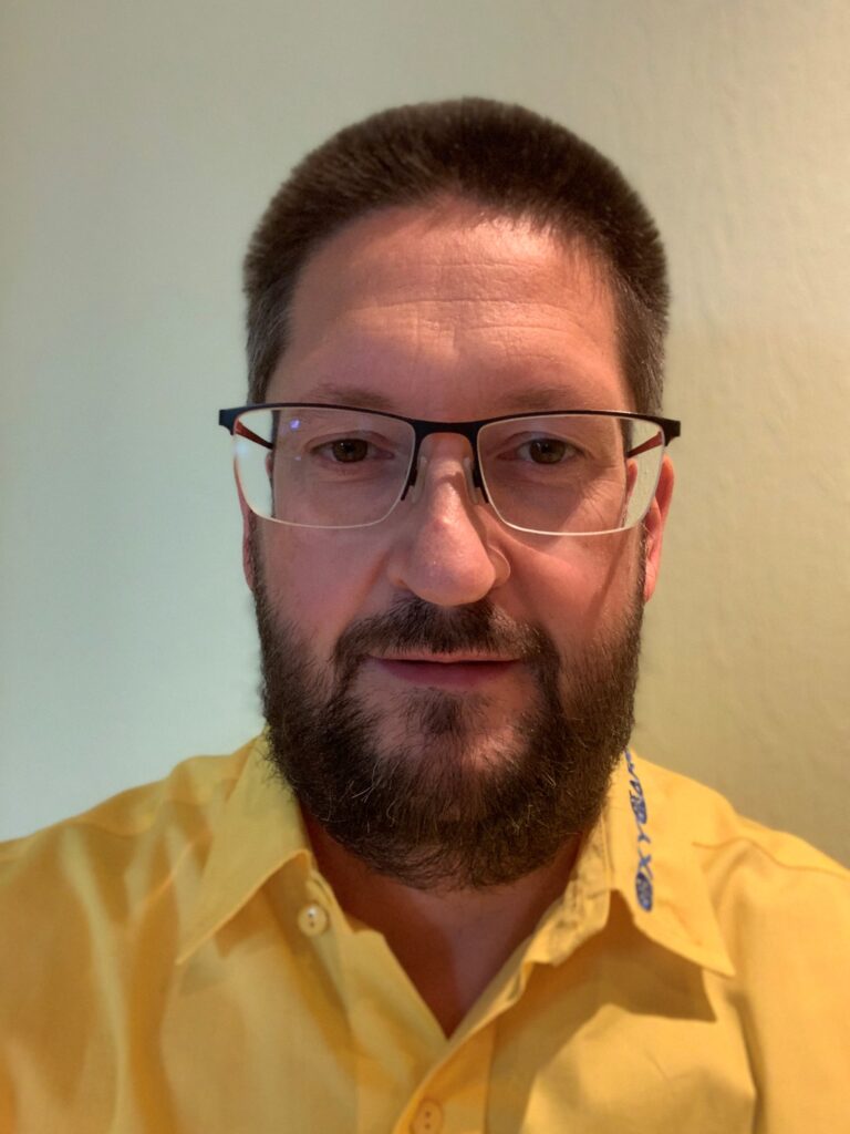 Foto von Thomas Sczuka, Mitarbeiter von OxyCare. Er trägt ein gelbes Hemd mit dem OxyCare Logo und schaut freundlich in die Kamera.